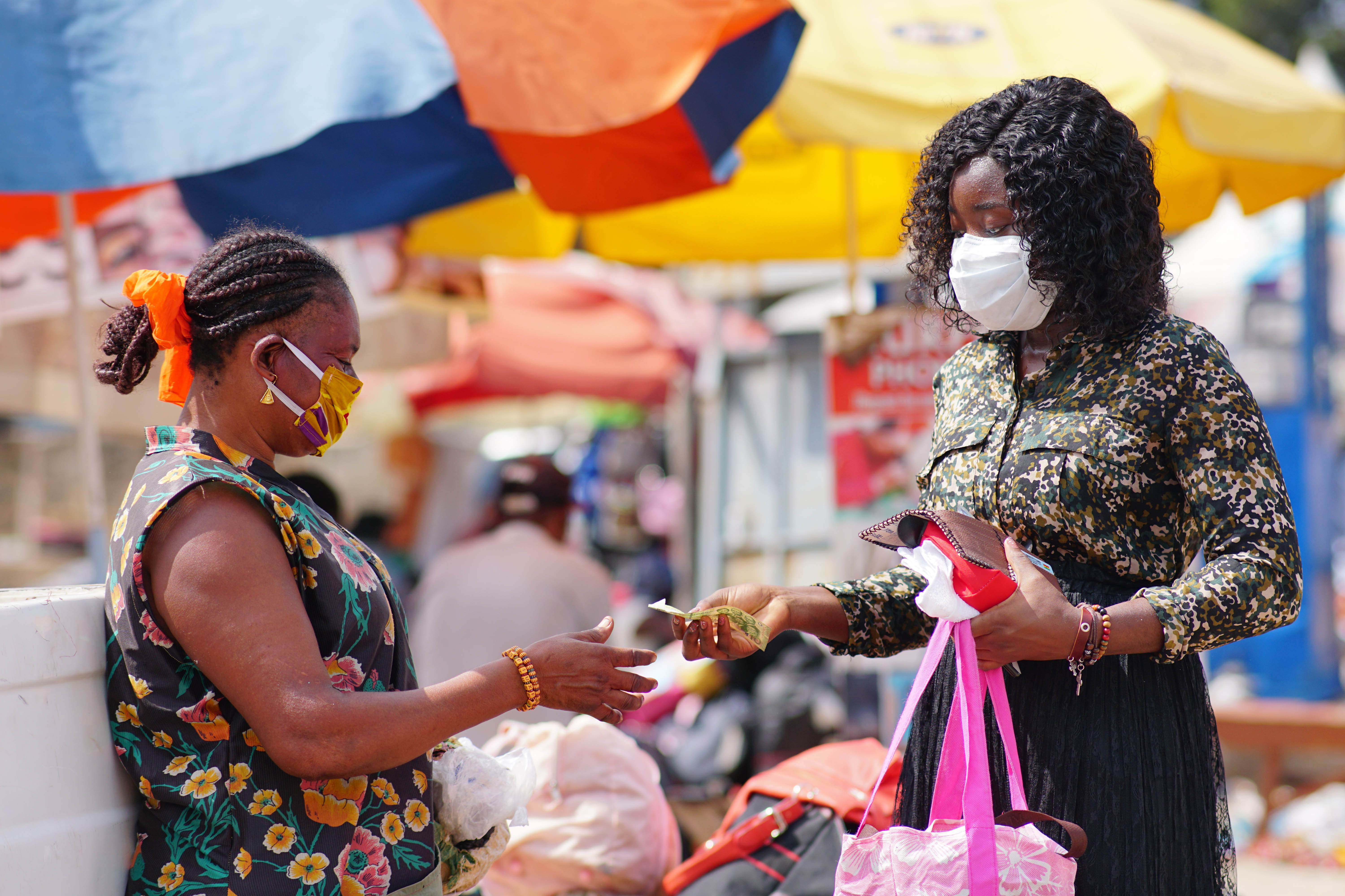 Young black woman paying money to a vendor for a purchased item in the market. Two women wearing locally made mask and surgical mask on the street.